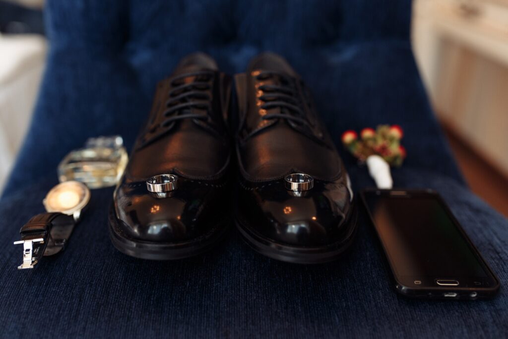 A pair of polished black dress shoes lies on a blue chair, surrounded by a watch, smartphone, and a small bouquet.