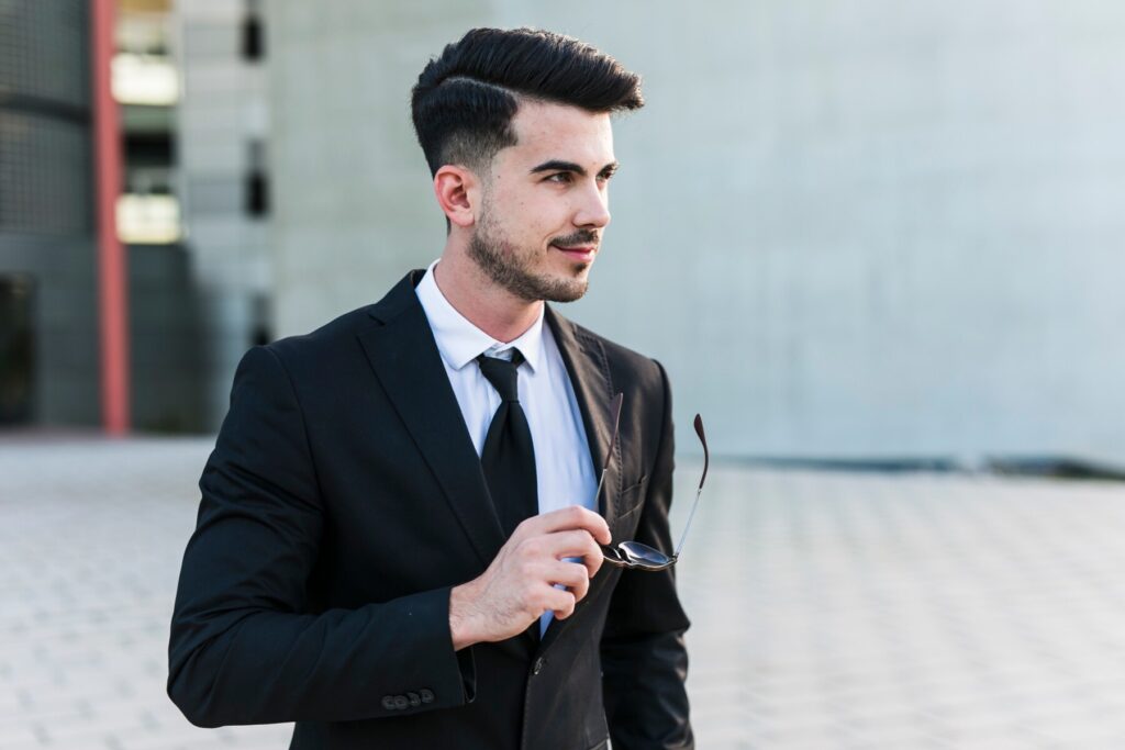 A man in a black suit holds sunglasses in one hand, standing against a minimalistic urban background with a gray wall.