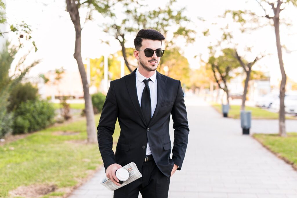 A man in a sleek black suit and tie walks confidently down a tree-lined path, holding a clutch purse.