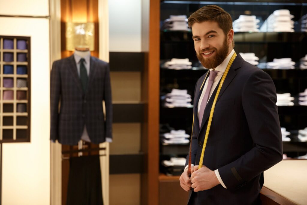 A man in a tailored suit holds a measuring tape in a high-end clothing store, showcasing neatly arranged shirts and a suit on display.
