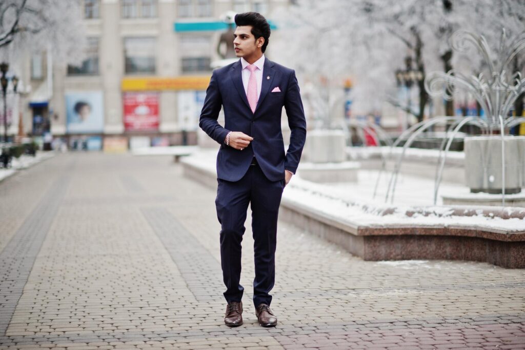 A well-dressed man in a navy suit and pink tie walks confidently through a snowy urban park, showcasing a stylish and polished look.