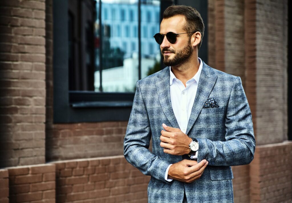 A well-dressed man in a stylish gray plaid blazer adjusts his watch, standing against a brick wall with a reflective window behind him.
