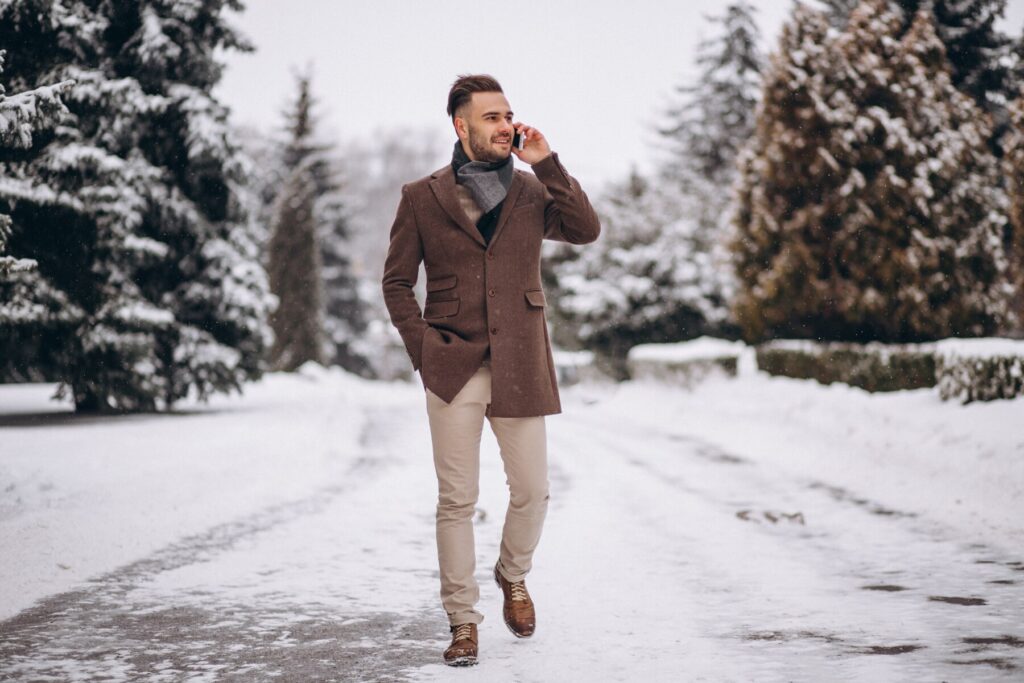 A man in a brown coat and scarf walks through a snow-covered path lined with trees, exuding a stylish winter vibe.