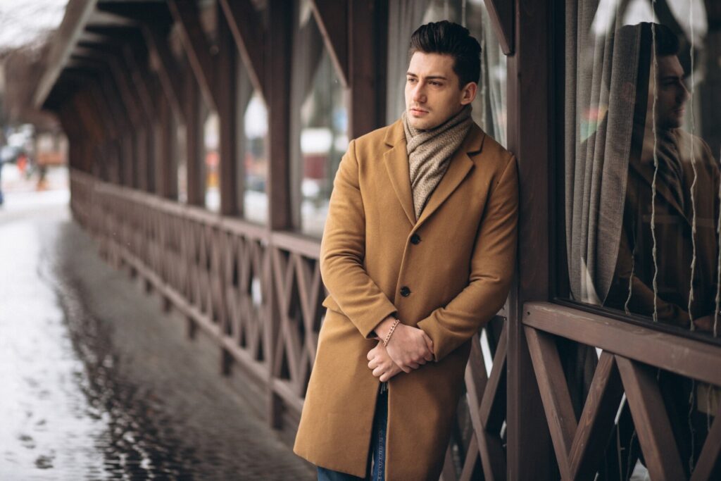A person in a stylish brown coat and a knitted scarf leans against a wooden railing, reflecting on a snowy street.
