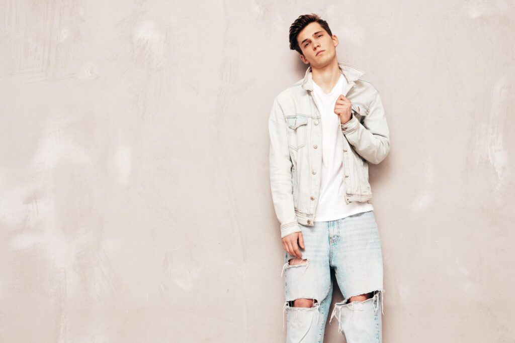A person stands against a textured light beige wall, wearing a denim jacket, a white shirt, and distressed light blue jeans.