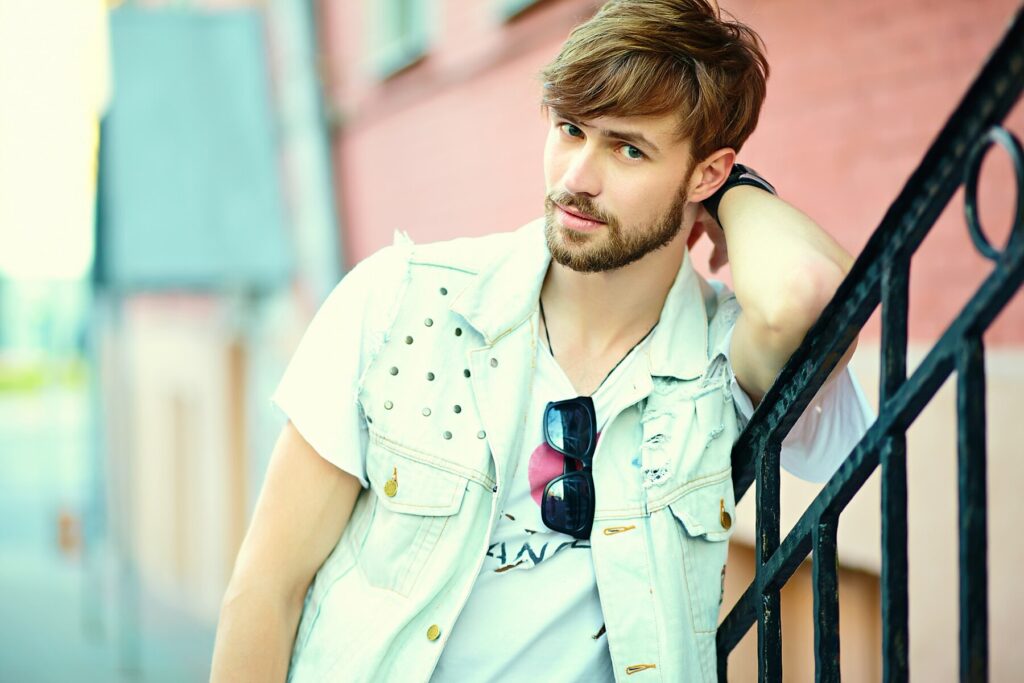 A young man leans against a railing, wearing a sleeveless denim vest over a graphic t-shirt and stylish sunglasses, in a city setting.