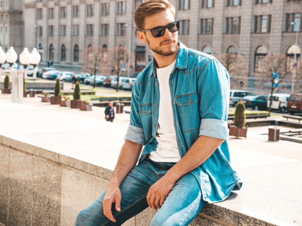 A young man sits casually on a stone ledge, wearing a denim shirt and jeans, with a city street and parked cars in the background.
