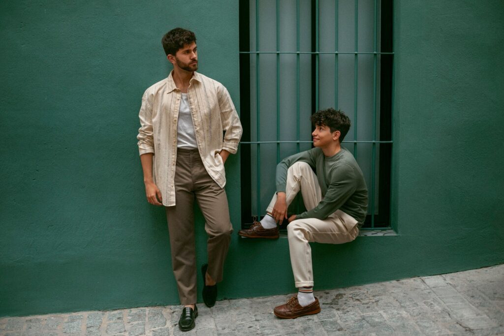 Two men dressed in casual attire pose against a green wall; one stands while the other sits casually by a window.