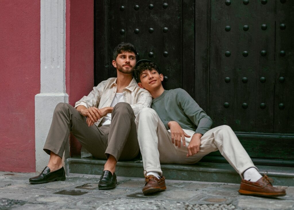 Two young men sit closely together on a cobblestone street, wearing casual outfits and leaning against a large, dark wooden door.