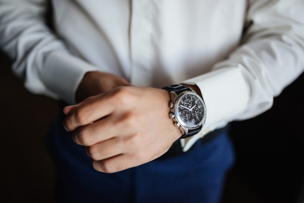 A close-up of a man's hand adjusting a stylish silver watch while wearing a white shirt and blue trousers.