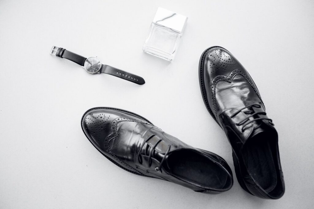 A pair of polished black dress shoes, a sleek timepiece, and a glass perfume bottle on a minimalistic surface.