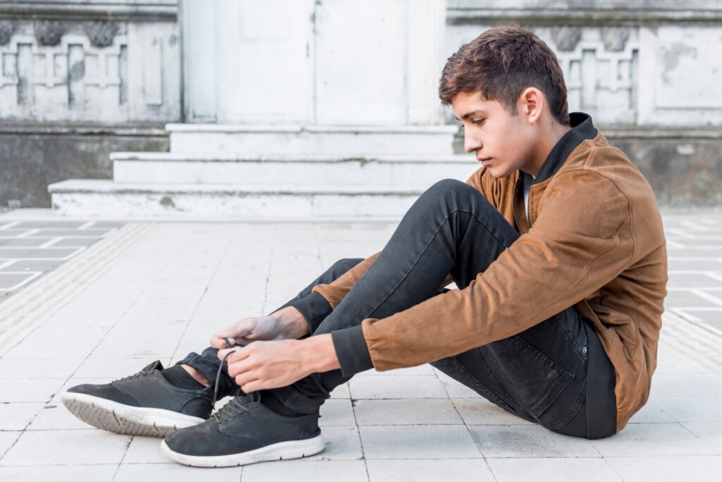 A person sits on tiled pavement, tying his black shoes. He's dressed in a brown jacket and black pants, with steps and a wall in the background.