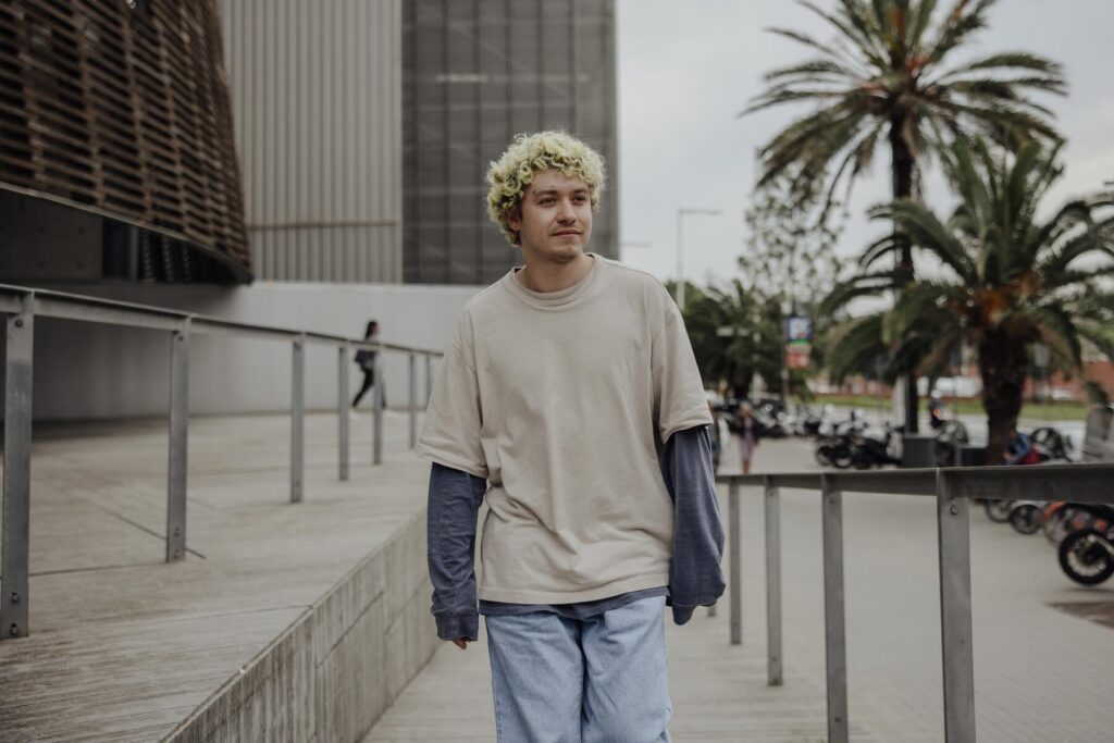 A person with curly blonde hair walks along a wooden pathway, surrounded by palm trees and modern architecture.