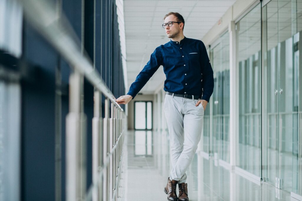 A man in a dark shirt and light pants leans against a railing in a modern hallway with glass walls and bright lighting.