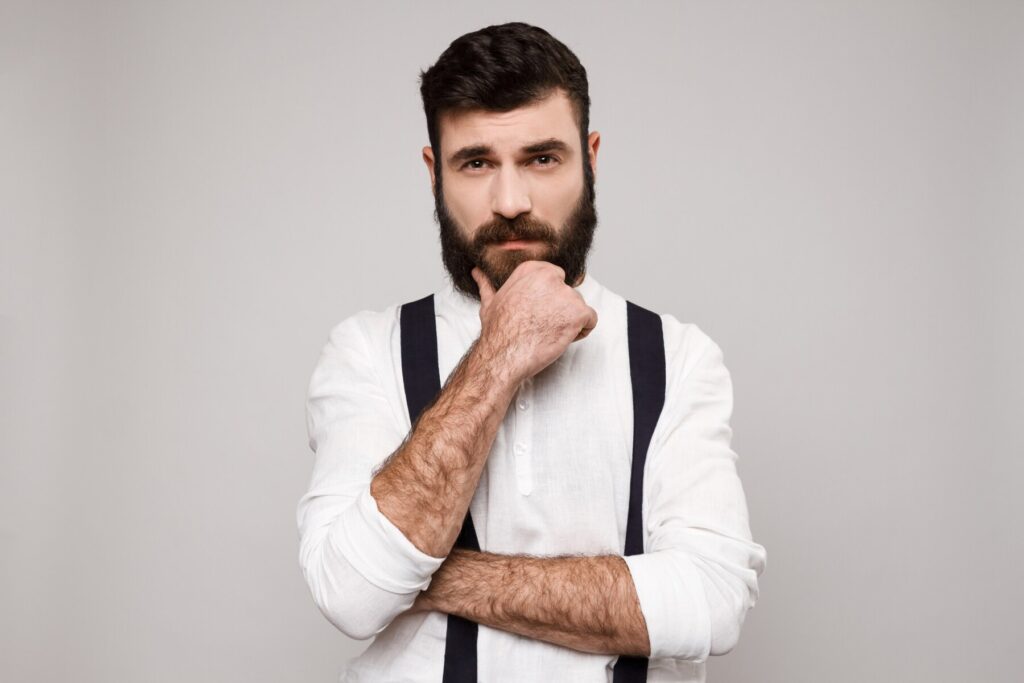 A man in a white shirt and black suspenders stands confidently with arms crossed, set against a neutral gray background.