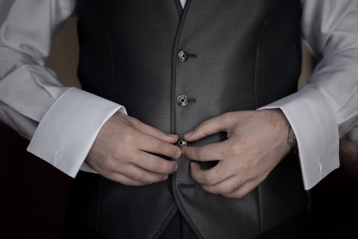 A man wearing a black vest over a white shirt, standing confidently with a neutral expression.
