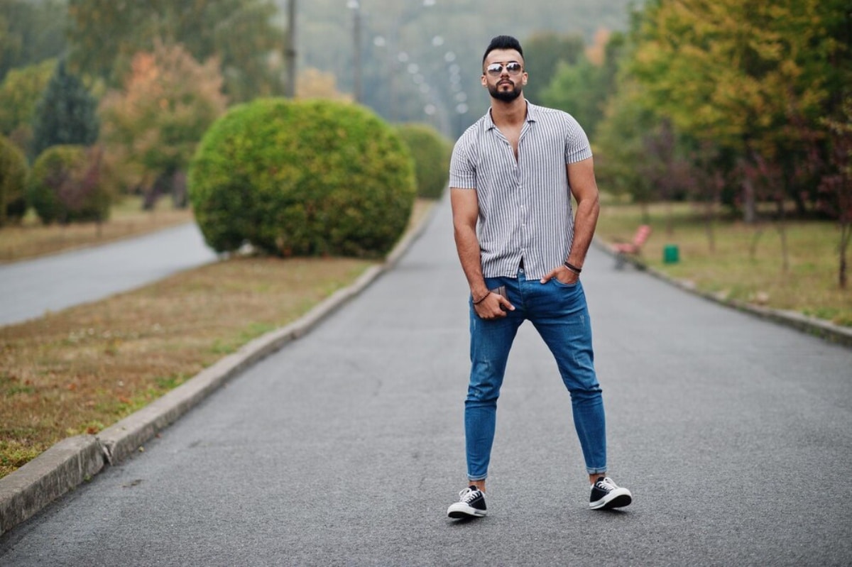 A man wearing sunglasses stands confidently on a road, surrounded by greenery and clear blue skies.