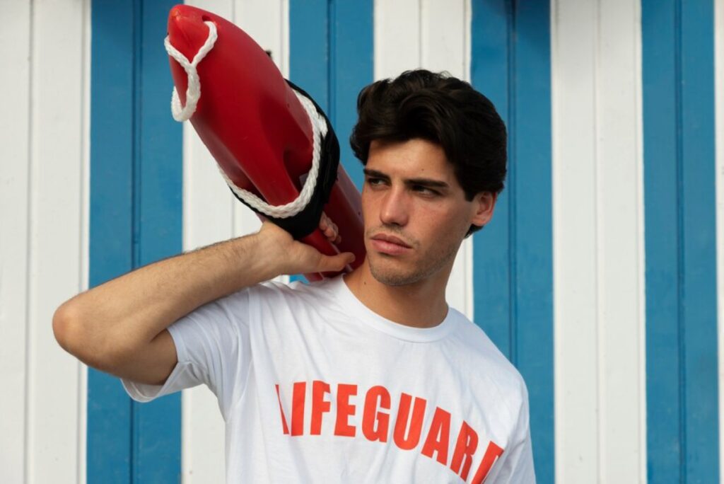 A man stands holding a bright red lifeguard helmet, ready for duty at the beach or poolside.