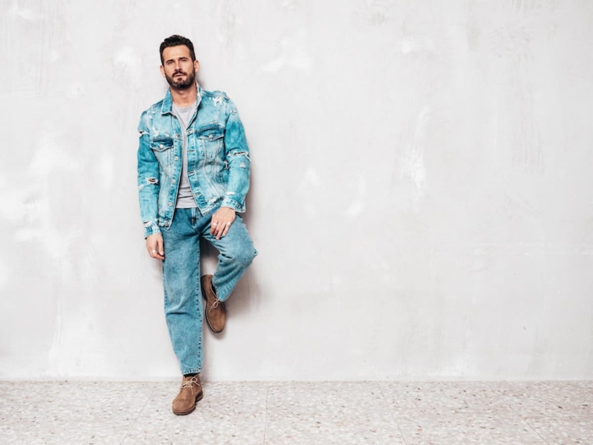 A man wearing jeans and a blue jacket stands casually against a textured wall.