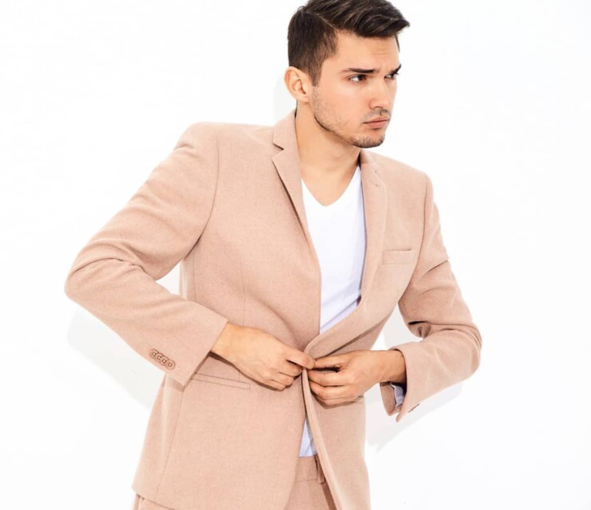 A man in a tan suit adjusts his jacket, standing confidently with a slight smile on his face.