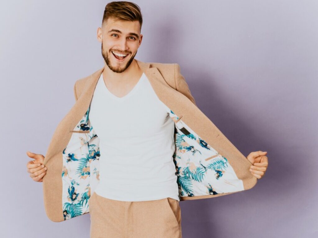 Young man wearing a tan suit and a floral patterned jacket, standing confidently with a neutral background.