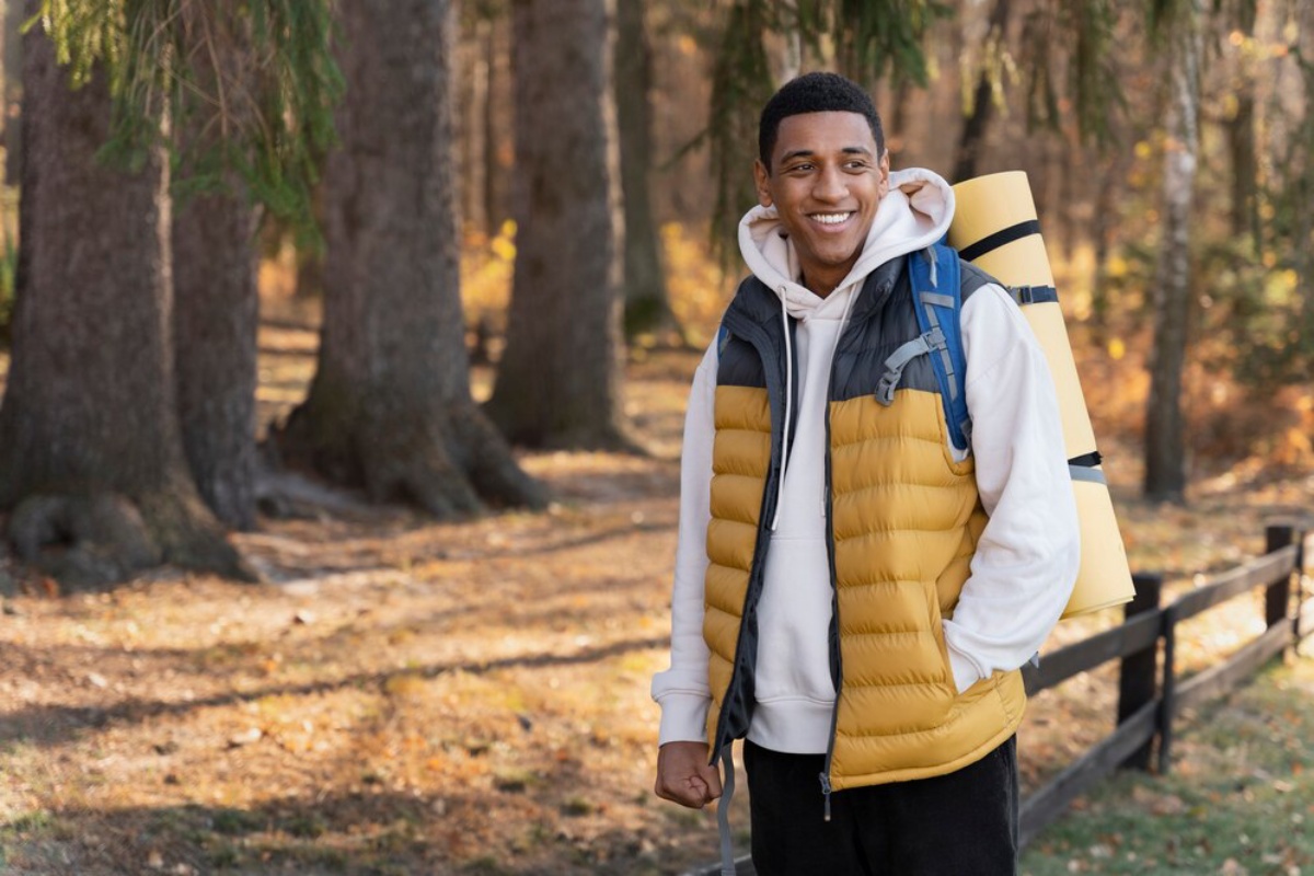  A young man wearing a yellow vest and carrying a backpack stands among trees in a wooded area.
