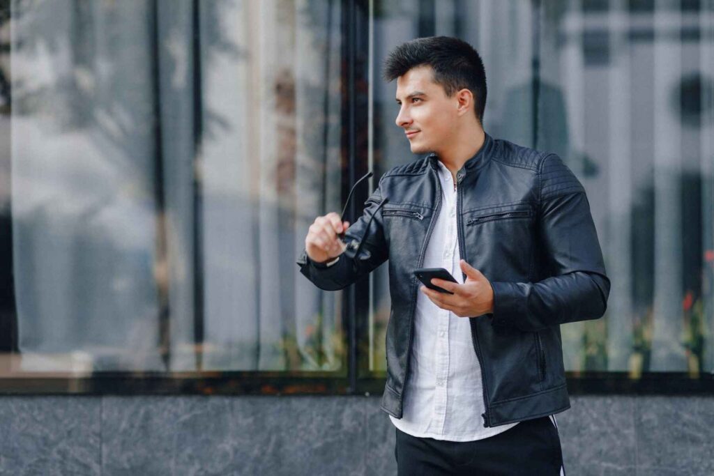 A man in a black leather jacket and white shirt holds sunglasses in one hand and a smartphone in the other, standing in front of large windows.